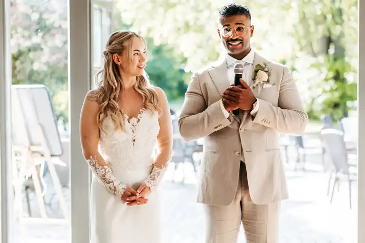 Eine Braut in einem weißen Spitzenkleid und ein Bräutigam in einem beigen Anzug stehen auf ihrer Hochzeit Gut am See. Der Bräutigam hält ein Mikrofon in der Hand und lächelt, während die Braut ihn anschaut. Beide wirken glücklich und werden von natürlichem Licht angestrahlt.