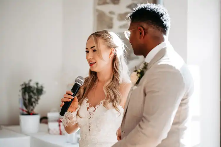 Eine Braut in einem weißen Spitzenkleid hält ein Mikrofon und spricht neben dem Bräutigam in einem hellen Anzug auf ihrer Hochzeit auf Gut am See. Sie stehen in einem Innenraum, in sanftes Sonnenlicht getaucht, vor einem weißen Hintergrund - aufgenommen von Hochzeitsfotograf Görlitz.