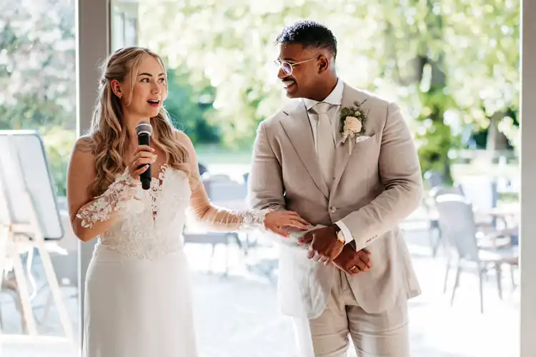 Eine Braut in einem weißen Kleid spricht in ein Mikrofon, während sie lächelt und die Hände des Bräutigams hält, der einen hellbeigen Anzug und eine Brille trägt. Aufgenommen von Hochzeitsfotograf Görlitz auf Hochzeit Gut am See, strömt das Sonnenlicht hinter den beiden in den Innenraum.