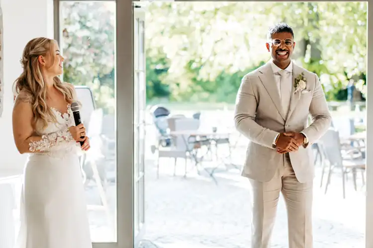 Eine lächelnde Braut, die ein Mikrofon in der Hand hält, blickt ihren Bräutigam in einem hellbeigen Anzug an, während das Sonnenlicht durch die offenen Türen hinter ihnen auf ihrer Hochzeit Gut am See strömt.