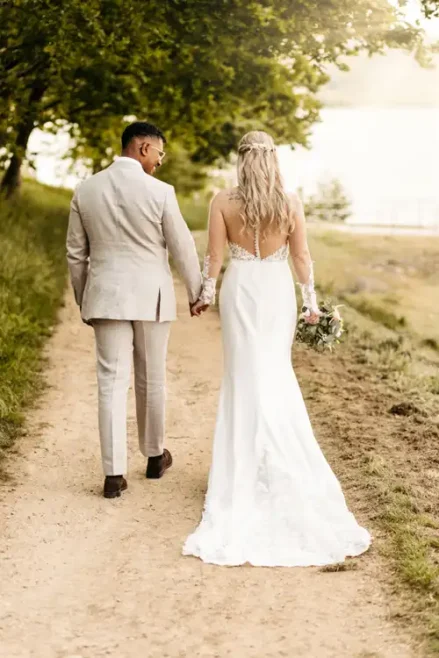 Eine Braut in einem weißen Kleid und ein Bräutigam in einem hellen Anzug gehen Hand in Hand einen von Bäumen gesäumten Weg auf Hochzeit Gut am See entlang, während die Braut einen Blumenstrauß hält und im Hintergrund ein See zu sehen ist.