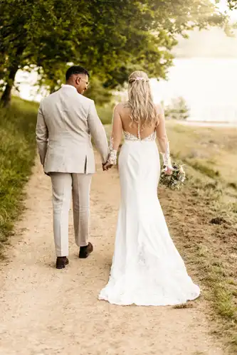 Eine Braut in einem weißen Kleid und ein Bräutigam in einem hellen Anzug gehen Hand in Hand einen von Bäumen gesäumten Weg auf Hochzeit Gut am See entlang, während die Braut einen Blumenstrauß hält und im Hintergrund ein See zu sehen ist.