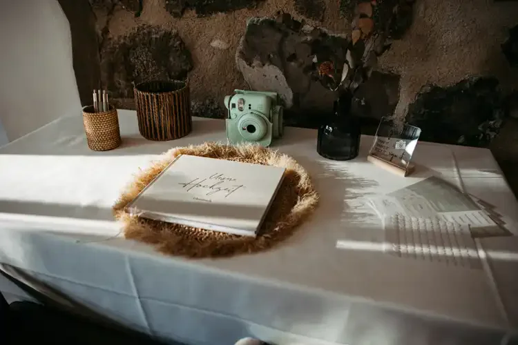 Ein Gästebuch auf einer weißen Tischdecke, umgeben von einer Strohmatte, einer grünen Sofortbildkamera, Trockenblumen in einer dunklen Vase, Stiften in einem Halter und Notizkarten - perfekt gestylt für eine Hochzeit Gut am See vor einer rustikalen Steinmauer.