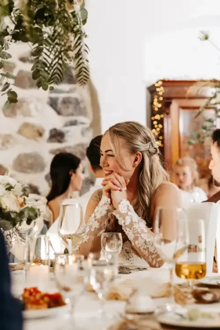 Eine Braut mit langen blonden Haaren in einem Spitzenhochzeitskleid lächelt und fasst sich an einem geschmückten Tisch, umgeben von Gästen und Blumenarrangements auf ihrem Hochzeitsempfang in Gut am See, in die Hände.