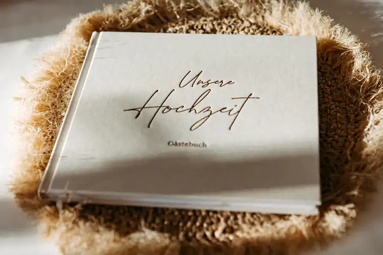 Ein beiges Hochzeitsgästebuch mit Unser Hochzeit Gästebuch in eleganter Schrift liegt auf einer strukturierten, gewebten Matte in warmem, natürlichem Licht - perfekt für die Feier Ihrer Hochzeit auf Gut am See.