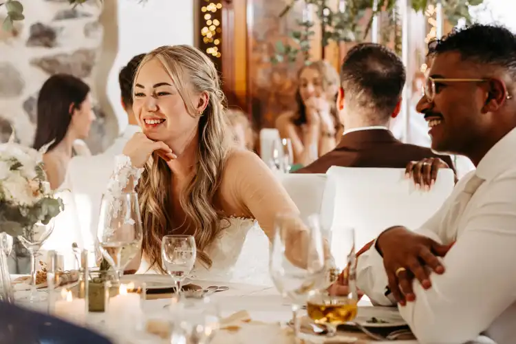 Eine lächelnde Braut mit langen blonden Haaren sitzt an einem geschmückten Tisch neben einem Mann mit Brille während einer Hochzeit auf Gut am See, wunderschön eingefangen von Hochzeitsfotograf Görlitz inmitten von Kerzen, Blumen und fröhlichen Gästen.