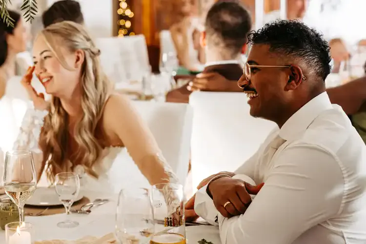 Eine lächelnde Braut und ein lächelnder Bräutigam sitzen während ihrer Hochzeit auf Gut am See zusammen an einem geschmückten Tisch, umgeben von Gästen. Beide wirken glücklich und sind in ein Gespräch vertieft, vor ihnen stehen Gläser und Tischgedecke.