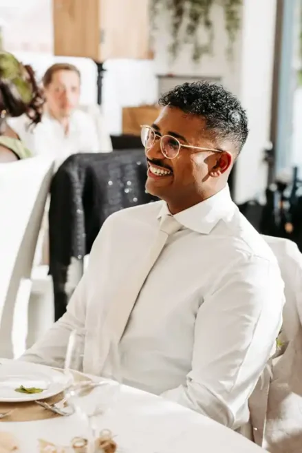 Ein Mann mit Brille, weißem Hemd und weißer Krawatte sitzt an einem Tisch und lächelt. Bei der hellen Indoor-Veranstaltung auf Hochzeit Gut am See mischen sich fröhliche Gäste im Hintergrund.