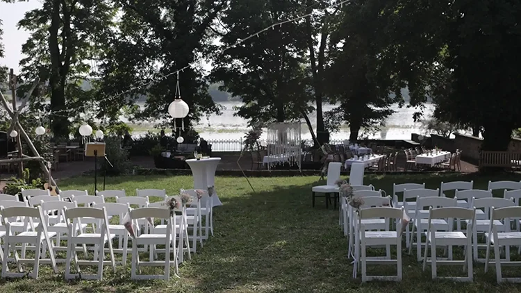 Eine Hochzeit im Freien besteht aus Reihen weißer Stühle vor einem Altar, umgeben von Bäumen. Weiße Laternen hängen über dem Sitzbereich. Im Hintergrund ist ein See zu sehen, der das Licht reflektiert. Ein Hochzeitsfotograf in Marcolinis Welt hält jeden bezaubernden Moment inmitten der mit weißen Tischdecken geschmückten Tische fest.