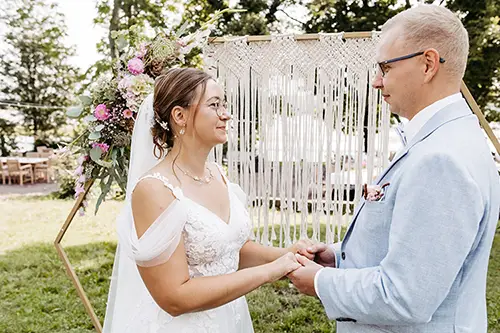 Braut und Bräutigam stehen sich im Freien Händchen haltend gegenüber. Die Braut trägt ein weißes Kleid mit Spitzendetails und Schleier. Der Bräutigam trägt einen hellgrauen Anzug. Dahinter befinden sich ein Makramee-Hintergrund im Boho-Stil und ein Blumenarrangement, wunderschön eingefangen von der Hochzeitsfotografin aus Dresden, Melanie Kunert Fotografie.