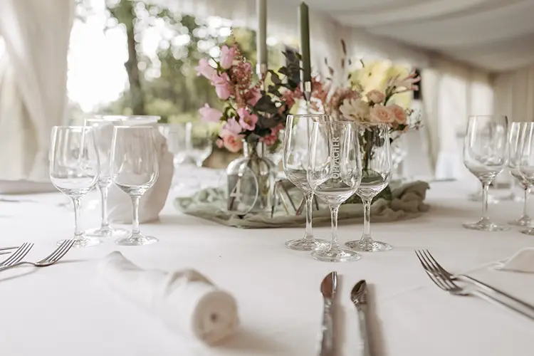 Ein wunderschön gedeckter Tisch mit elegantem Glasgeschirr, einem Blumen- und Grünschmuck sowie hohen, schlanken Kerzen erinnert an die Hochzeit in Marcolinis Welt. Im Hintergrund spiegelt sich ein weiches, weiß drapiertes Zelt und zwei gerahmte Spiegel, die die Atmosphäre widerspiegeln.