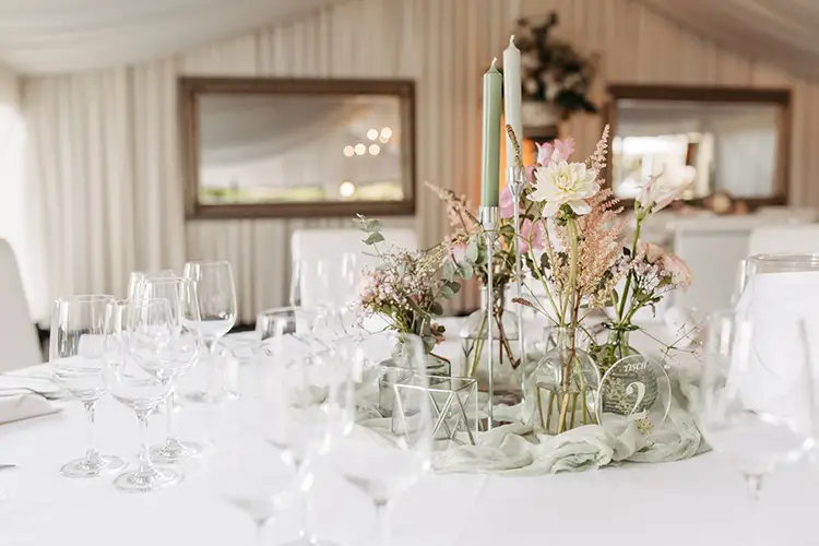 Ein wunderschön gedeckter Tisch mit elegantem Glasgeschirr, einem Blumen- und Grünschmuck sowie hohen, schlanken Kerzen erinnert an die Hochzeit in Marcolinis Welt. Im Hintergrund spiegelt sich ein weiches, weiß drapiertes Zelt und zwei gerahmte Spiegel, die die Atmosphäre widerspiegeln.
