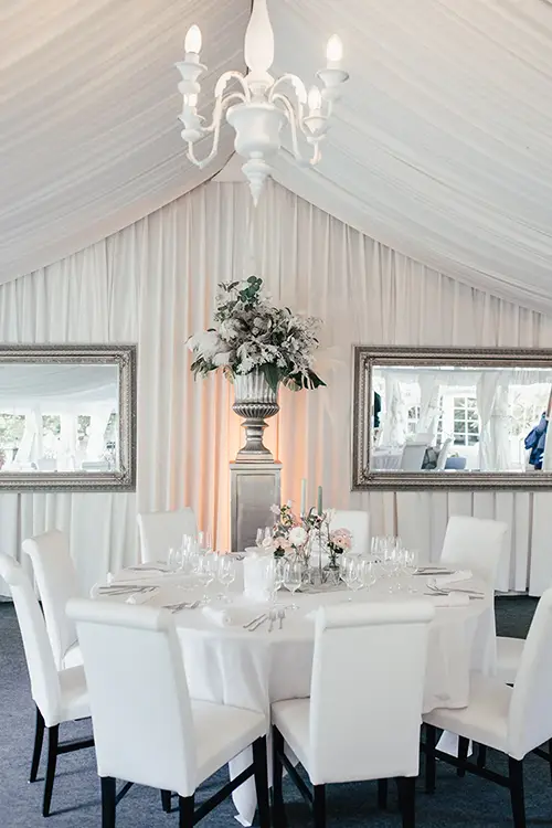 Ein eleganter, weißer Essbereich spiegelt den Charme der Hochzeit Marcolinis Welt Dresden wider. Der Kronleuchter erstrahlt in einem warmen Licht über dem runden Tisch mit weißen Stühlen. Zwei große Spiegel und ein florales Tischdekor unterstreichen das elegante Ambiente – ideal für Hochzeitsfotografen aus Dresden.