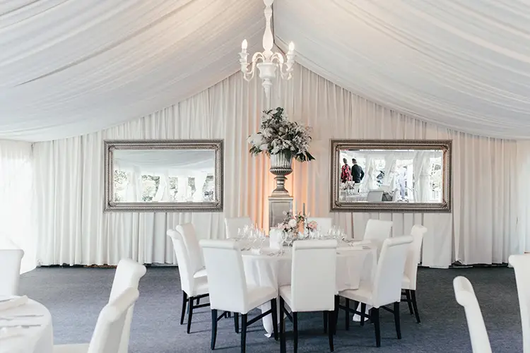 Ein eleganter, weißer Essbereich spiegelt den Charme der Hochzeit Marcolinis Welt Dresden wider. Der Kronleuchter erstrahlt in einem warmen Licht über dem runden Tisch mit weißen Stühlen. Zwei große Spiegel und ein florales Tischdekor unterstreichen das elegante Ambiente – ideal für Hochzeitsfotografen aus Dresden.
