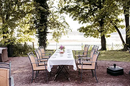 Ein langer, rechteckiger Gartentisch mit weißer Tischdecke wird von zehn Korbstühlen flankiert und spiegelt perfekt den Geist der Hochzeit in Marcolinis Welt wider. Er steht auf einem Kiesweg unter hohen Bäumen, in der Mitte steht eine Vase mit rosa Blumen.