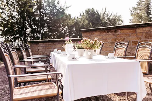 Inmitten der bezaubernden Kulisse, die an „Hochzeit in Marcolinis Welt“ erinnert, ist ein langer Esstisch im Freien mit einer weißen Tischdecke, eleganten weißen Tassen und Untertassen sowie Blumenarrangements in makellosen Vasen geschmückt. Holzstühle umgeben den Tisch, eingerahmt von einer rustikalen Steinmauer und sonnendurchfluteten Bäumen.