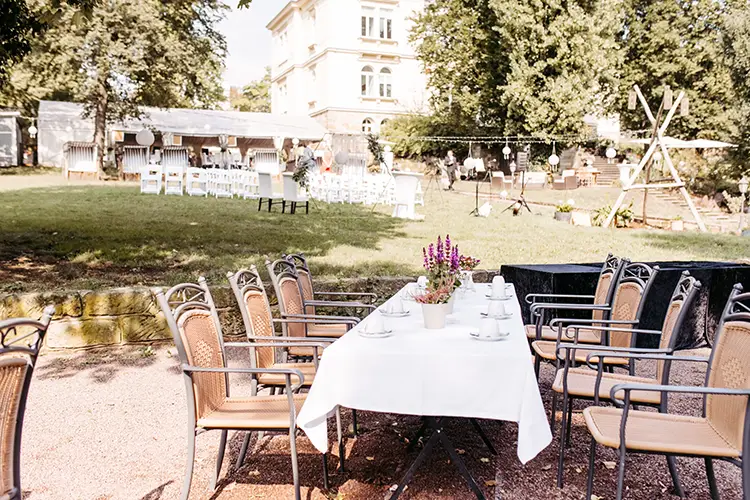 Eine bezaubernde Gartenanlage im Marcolinis Welt ist Schauplatz einer Hochzeit. Die Tische sind mit weißen Tischdecken bedeckt und mit Blumentöpfen geschmückt. Im Hintergrund zieren weitere Sitzgelegenheiten die Rasenfläche, geschützt von Bäumen und einem stattlichen Gebäude in der Nähe.