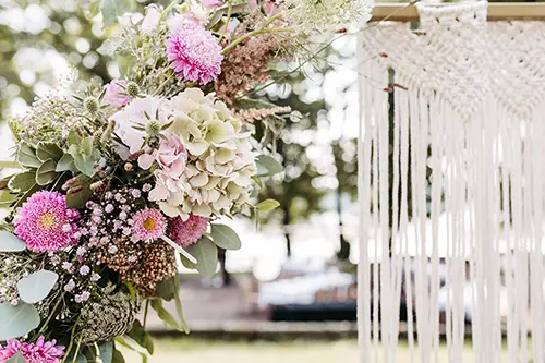 Nahaufnahme eines Blumenarrangements mit rosa und weißen Blumen, Grünpflanzen und strukturierten Pflanzen. Rechts davon sorgt ein Makramee-Behang für eine sanfte, unkonventionelle Atmosphäre. Melanie Kunert Fotografie hat diese Schönheit in Hochzeit Marcolinis Welt Dresden vor unscharfem Grün und Bokeh-Licht eingefangen.