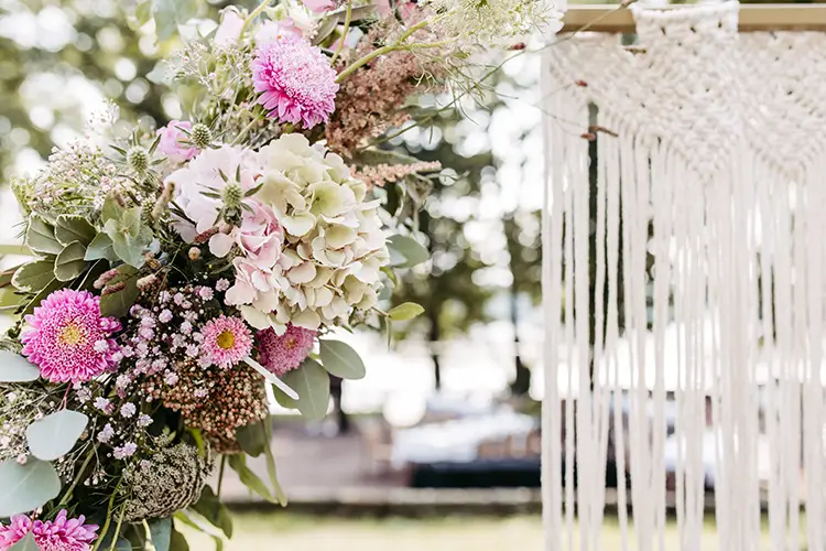 Nahaufnahme eines Blumenarrangements mit rosa und weißen Blumen, Grünpflanzen und strukturierten Pflanzen. Rechts davon sorgt ein Makramee-Behang für eine sanfte, unkonventionelle Atmosphäre. Melanie Kunert Fotografie hat diese Schönheit in Hochzeit Marcolinis Welt Dresden vor unscharfem Grün und Bokeh-Licht eingefangen.