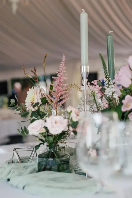 Ein elegant dekorierter Tisch mit hohen grünen Kerzen, rosa und weißen Blumen in Vasen und einer zartgrünen Tischdecke. Gläser verschwimmen im Vordergrund und schaffen eine elegante und einladende Atmosphäre, die an eine Hochzeit in Marcolinis Welt erinnert.