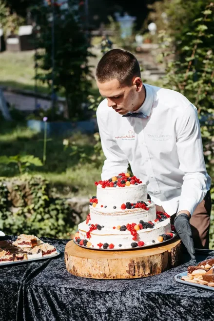 Eine Person im weißen Hemd dekoriert eine mehrstöckige Hochzeitstorte mit Beeren, was an eine Hochzeit in Marcolinis Welt erinnert. Die Torte steht auf einem Holzsockel auf einer dunklen Tischdecke, umgeben von Tellern mit geschnittener Torte und üppigem Grün im Hintergrund.