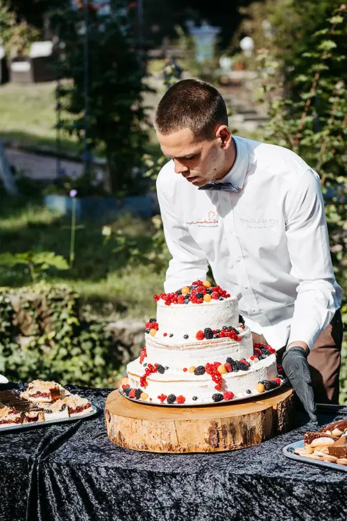 Eine Person im weißen Hemd dekoriert eine mehrstöckige Hochzeitstorte mit Beeren, was an eine Hochzeit in Marcolinis Welt erinnert. Die Torte steht auf einem Holzsockel auf einer dunklen Tischdecke, umgeben von Tellern mit geschnittener Torte und üppigem Grün im Hintergrund.