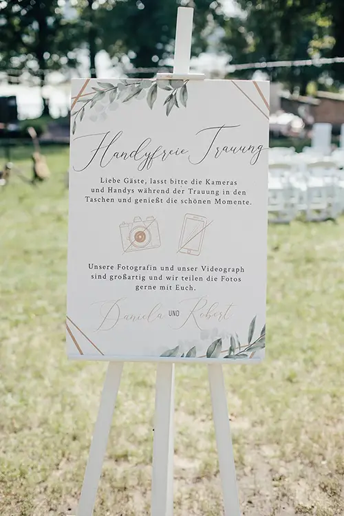 Ein mit Blumenmotiven verziertes Schild bei der Hochzeit in Marcolinis Welt bittet die Gäste, Handys und Kameras wegzulegen und den Moment zu genießen. So können Fotograf und Videofilmer wunderschöne Fotos teilen.