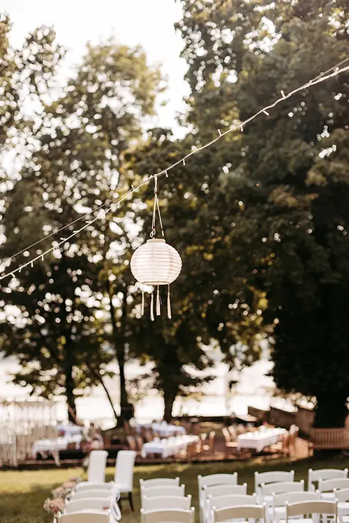 Hochzeitsaufbau im Freien mit weißen Stühlen vor einem Altar, der die Eleganz einer Hochzeit in Marcolinis Welt einfängt. Eine Lichterkette und eine Papierlaterne hängen darüber, während Bäume eine natürliche Kulisse bilden. Im Hintergrund sind Tische im gesprenkelten Sonnenlicht aufgestellt.