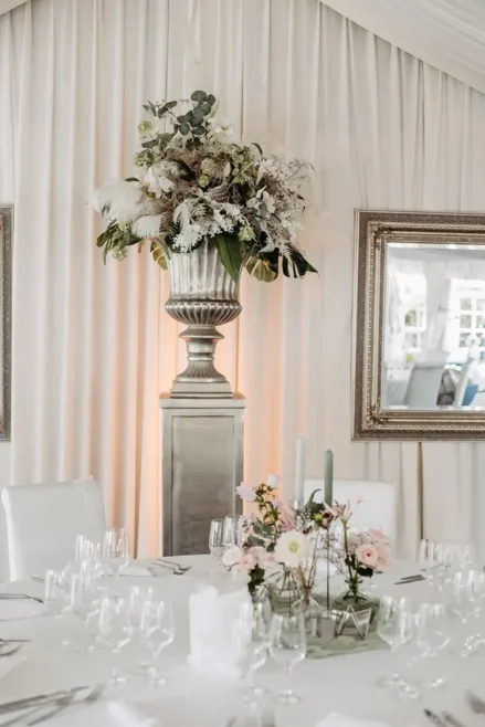 Elegantes Event-Setup, das an eine Hochzeit in Marcolinis Welt erinnert: Ein runder weißer Tisch, geschmückt mit rosa und weißen Blumen und grünen Kerzen. Ein großes Blumenarrangement auf einem Podest ziert die Szene, kontrastiert mit drapiertem Stoff und gerahmten Spiegeln an der Wand.
