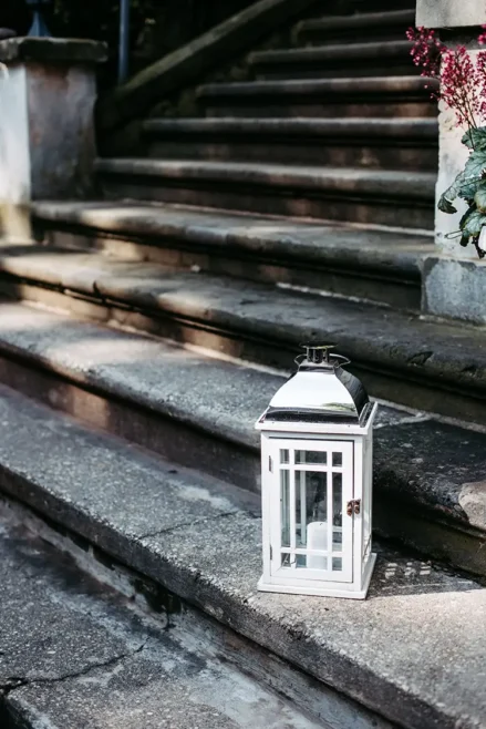 Eine weiße Laterne mit Glaseinsätzen steht auf den abgenutzten Steinstufen und wird teilweise vom Sonnenlicht beleuchtet. Diese verwitterten Stufen führen nach oben, als würden sie zu einer Hochzeit in Marcolinis Welt führen, während rechts ein Strauß kleiner roter Blumen für einen Hauch von lebendiger Farbe sorgt.