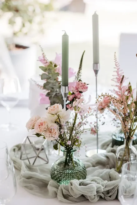 Eine filigrane Tischdekoration für eine Hochzeit in Marcolinis Welt besteht aus grünen Kerzen, rosa Blumen in strukturierten Glasvasen und üppigem Grün auf grauem Stoff. Das Arrangement umfasst geometrisches Dekor, elegant auf einer weißen Tischdecke platziert.