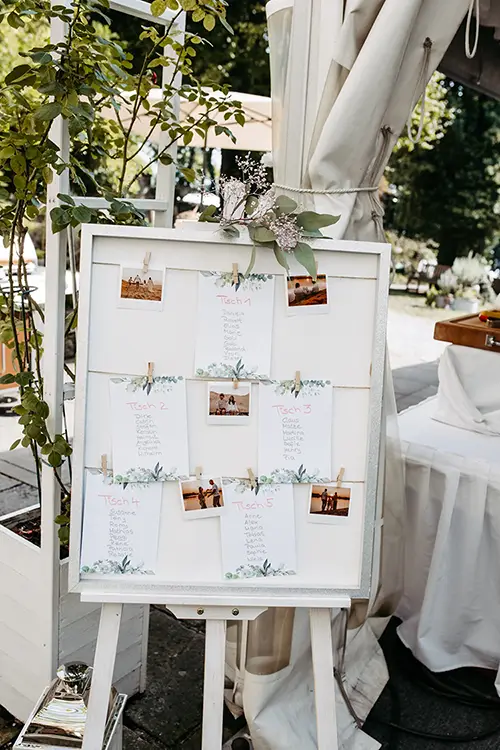 Der Sitzplan zum Thema „Hochzeit in Marcolinis Welt“ ruht elegant auf einer Staffelei und präsentiert Tischlisten, die mit dekorativem Laub und kleinen Fotos geschmückt sind. Im Freien platziert, besticht er durch üppiges Grün im Hintergrund und einen Blick auf ein weißes Baldachin darüber.