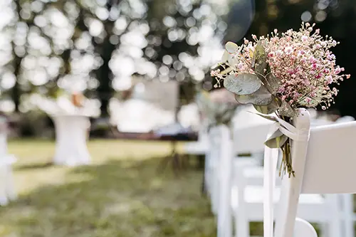 Ein zarter Strauß rosa Blumen und Grünpflanzen schmückt die Rückenlehne eines weißen Stuhls und fängt die Atmosphäre einer Hochzeit in Marcolinis Welt ein. Die Stühle sind im Freien angeordnet, mit verschwommenen Bäumen und Tischen im Hintergrund, was eine romantische Hochzeit oder Veranstaltung suggeriert.