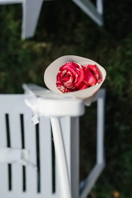 Eine einzelne rote Rose, in weißes Papier gewickelt, liegt auf einem weißen Stuhl. Die umgebende Rasenfläche und der Stuhl sind Teil eines gepflegten Arrangements, das an eine formelle Veranstaltung oder Zeremonie im Freien erinnert.