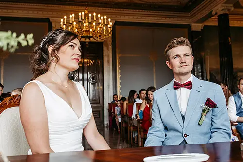 Im eleganten Schloss Albrechtsberg in Dresden sitzen eine Braut im weißen Kleid und ein Bräutigam im hellblauen Anzug mit roter Fliege an einem Holztisch. Im Hintergrund sind Gäste zu sehen, und ein kunstvoller Kronleuchter hängt von der Decke – die perfekte Kulisse für die Hochzeit.