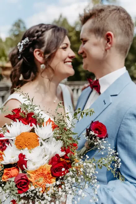 Ein fröhliches Paar im Hochzeitskleid blickt sich auf Schloss Albrechtsberg in Dresden an. Die Braut hält einen leuchtenden Strauß roter, weißer und orangefarbener Blumen mit Grünpflanzen in den Händen, während der Bräutigam in hellblauem Anzug und roter Fliege herzlich lächelt. Im unscharfen Hintergrund sind Bäume zu sehen.