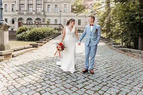 Eine Braut im weißen Kleid und ein Bräutigam im hellgrauen Anzug gehen Hand in Hand über einen Kopfsteinpflasterweg am Schloss Albrechtsberg, mit dem historischen Gebäude im Hintergrund. Sie lächeln sich an, im Sonnenlicht getaucht und umgeben von Dresdens üppigem Grün.