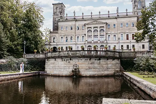Ein Paar steht an einem Teich vor dem prachtvollen Schloss Albrechtsberg in Dresden. Die kunstvolle Architektur, wunderschön eingefangen von Melanie Kunert Fotografie, besticht durch dekorative Balkone und hohe Türme. Üppiges Grün und ein leicht bewölkter Himmel runden die romantische Szene ab.