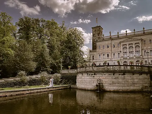 Ein Paar steht an einem Teich vor dem prachtvollen Schloss Albrechtsberg in Dresden. Die kunstvolle Architektur, wunderschön eingefangen von Melanie Kunert Fotografie, besticht durch dekorative Balkone und hohe Türme. Üppiges Grün und ein leicht bewölkter Himmel runden die romantische Szene ab.