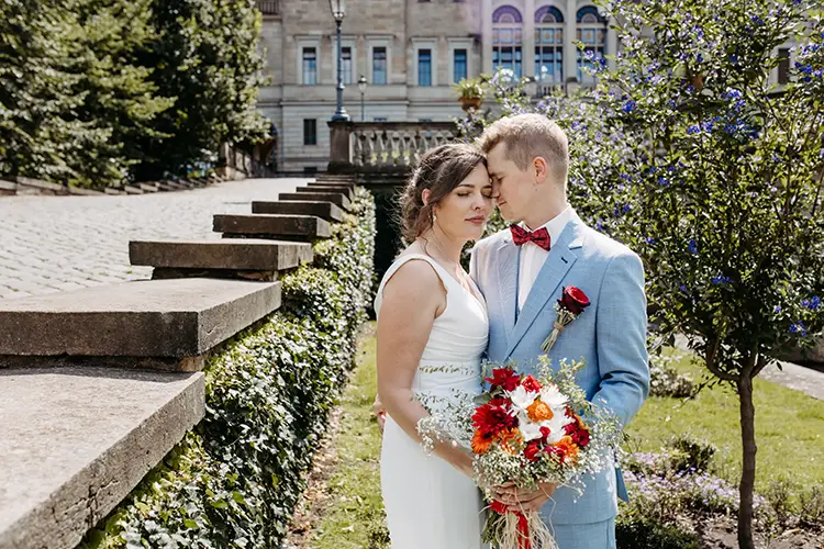 Braut und Bräutigam stehen eng beieinander auf einem Kopfsteinpflasterweg vor Schloss Albrechtsberg in Dresden. Die Braut trägt ein weißes Kleid und hält einen Strauß roter Blumen in der Hand, während der Bräutigam einen hellblauen Anzug mit roter Fliege trägt. Ein historisches Gebäude und üppiges Grün bilden die perfekte Kulisse für ihre Hochzeit.