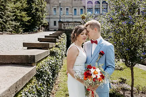 Braut und Bräutigam stehen eng beieinander auf einem Kopfsteinpflasterweg vor Schloss Albrechtsberg in Dresden. Die Braut trägt ein weißes Kleid und hält einen Strauß roter Blumen in der Hand, während der Bräutigam einen hellblauen Anzug mit roter Fliege trägt. Ein historisches Gebäude und üppiges Grün bilden die perfekte Kulisse für ihre Hochzeit.