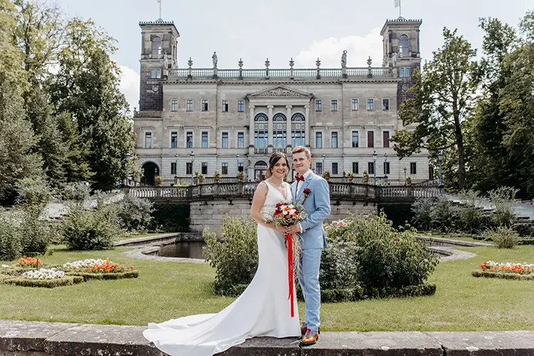 Im üppigen Garten von Schloss Albrechtsberg stehen eine Braut im weißen Kleid und ihr Bräutigam im hellgrauen Anzug nebeneinander. Die Sonne scheint hell am klaren Dresdner Himmel, und vor der Kulisse des kunstvollen Schlosses entfaltet sich diese atemberaubende Hochzeitsszene.