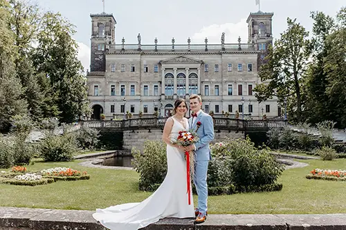 Im üppigen Garten von Schloss Albrechtsberg stehen eine Braut im weißen Kleid und ihr Bräutigam im hellgrauen Anzug nebeneinander. Die Sonne scheint hell am klaren Dresdner Himmel, und vor der Kulisse des kunstvollen Schlosses entfaltet sich diese atemberaubende Hochzeitsszene.