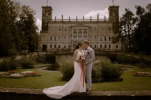 Eine Braut im weißen Kleid und ein Bräutigam im grauen Anzug stehen eng beieinander und halten Händchen im eleganten Schloss Albrechtsberg Dresden. Eingefangen von Melanie Kunert Fotografie, sind sie umgeben von üppigem Grün und Blumen und schaffen so eine idyllisch-romantische Kulisse.