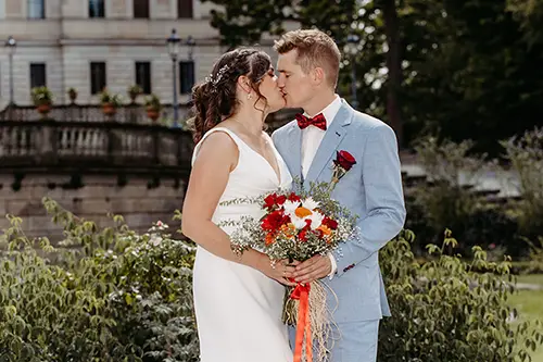 Braut und Bräutigam küssen sich im Freien vor Schloss Albrechtsberg in Dresden. Die Braut trägt ein weißes Kleid und hält einen Strauß roter, weißer und orangefarbener Blumen in der Hand, während der Bräutigam einen hellblauen Anzug mit roter Fliege trägt. Ein historisches Gebäude und üppiges Grün bilden die perfekte Kulisse für ihre Hochzeit.