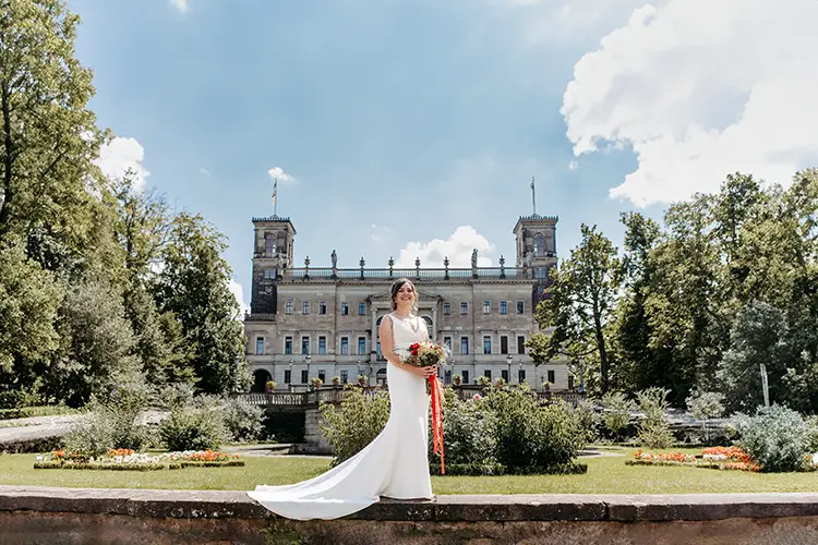 Eine Braut in weißem Kleid und Blumenstrauß posiert im Garten von Schloss Albrechtsberg Dresden, eingerahmt von seinen kunstvollen Türmen. Der leicht bewölkte Himmel wirft sanftes Licht auf das üppige Grün, wunderschön eingefangen von Hochzeitsfotografin Dresden – Melanie Kunert Fotografie.
