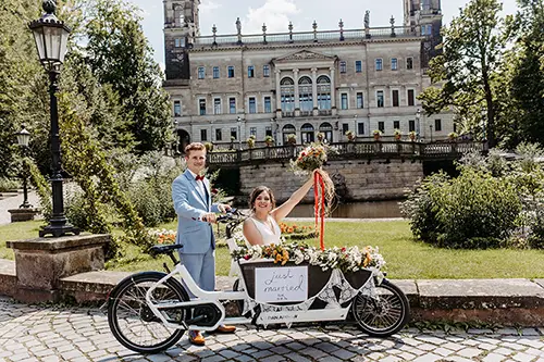 Braut und Bräutigam posieren mit einem dekorierten Tandem mit der Aufschrift „Just Married“ vor dem prachtvollen Schloss Albrechtsberg in Dresden. Die Braut hält einen Blumenstrauß, während der Bräutigam neben ihr steht. Beide lächeln inmitten eines üppigen Gartens. Eingefangen von Melanie Kunert Fotografie.
