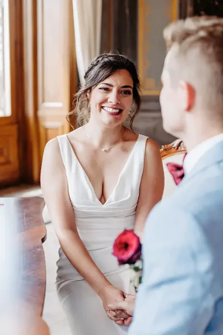 Eine lächelnde Braut im weißen Kleid sitzt ihrem Bräutigam im hellblauen Anzug auf Schloss Albrechtsberg in Dresden gegenüber. Sie hält rote Rosen in den Händen, wunderschön eingefangen von Melanie Kunert Fotografie. Das Paar befindet sich in einem holzgetäfelten Raum mit natürlichem Licht, das durch ein Fenster fällt.