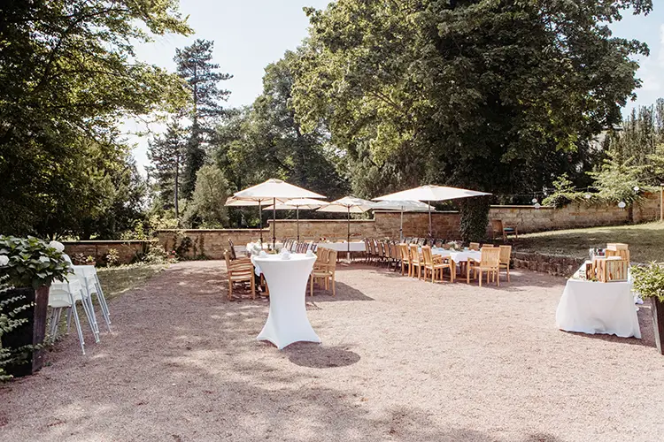 Outdoor-Event-Setup im sonnigen Garten in Dresden mit Holztischen und Stühlen unter großen Sonnenschirmen. Im Vordergrund steht ein hoher Cocktailtisch mit weißem Tuch. Üppige Bäume und eine Steinmauer umgeben den Bereich und schaffen eine ruhige Atmosphäre, perfekt für eine Hochzeit.