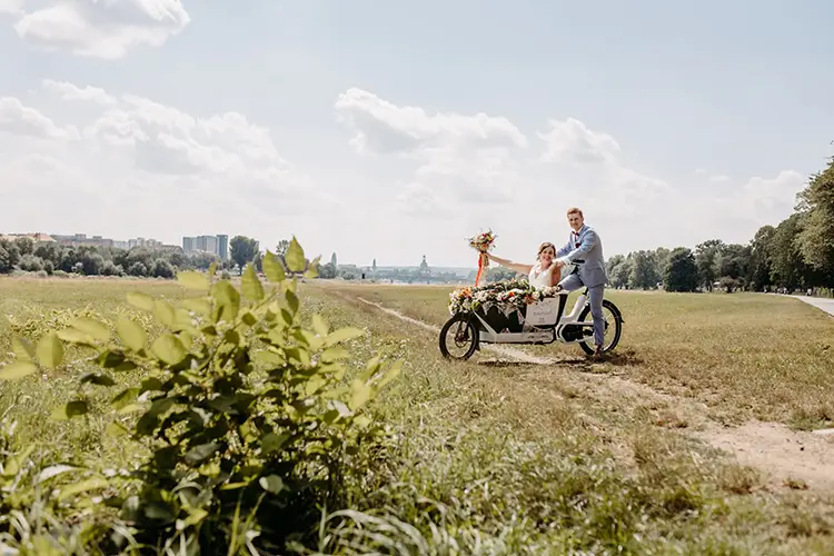 Ein Paar fährt mit einem blumengeschmückten Fahrrad über eine Wiese. Der Bräutigam im grauen Anzug tritt in die Pedale, während die Braut einen Blumenstrauß im Blumenkorb hält. Es ist sonnig, und in der Ferne ist zwischen Bäumen und Gebäuden das Dresdner Schloss Albrechtsberg zu sehen – eine idyllische Hochzeitskulisse.